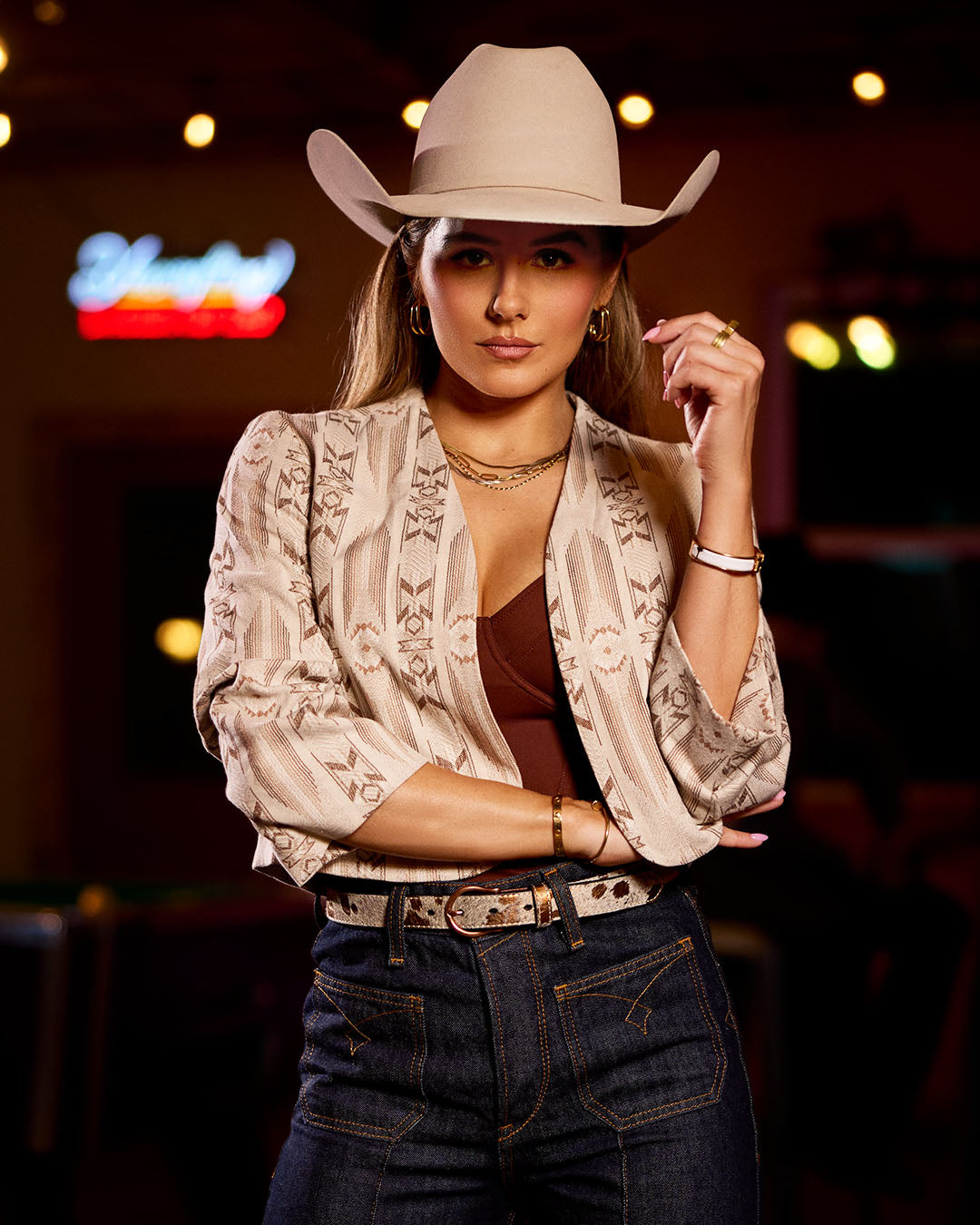 Woman wearing a cowboy hat and patterned jacket in a dimly lit room with neon signs.