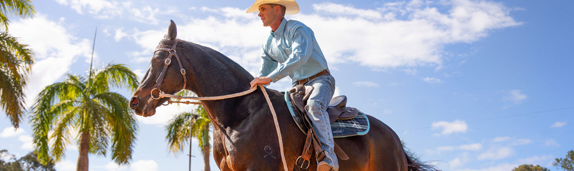 Cowboy riding brown horse wearing Ariat clothing and boots with blue sky and palm trees background.