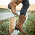 Person wearing brown cowboy boots with intricate designs, standing on a wooden fence.