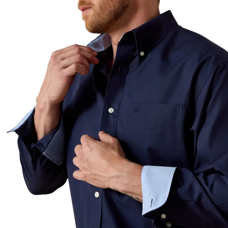 Man adjusting his navy blue shirt with light blue cuffs against a beige background
