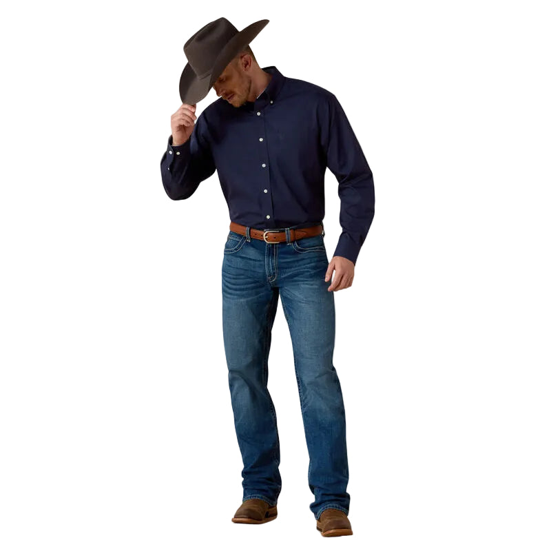Man wearing a navy shirt, blue jeans, and a brown hat against a beige background