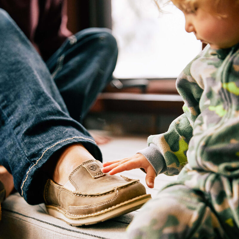 Child touching a brown shoe worn by an adult, with a blurred indoor background