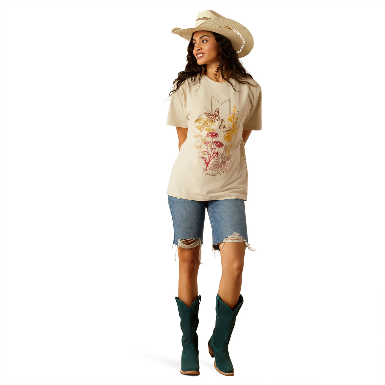 Woman wearing a beige t-shirt with floral and deer design, blue jeans, and a cowboy hat on a white background
