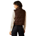 Person wearing a brown puffer vest over a cream shirt with a white background