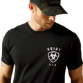 Man wearing a black t-shirt with Ariat logo and a camouflage cap on a white background