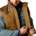 Man wearing a brown quilted vest over a denim jacket on a white background