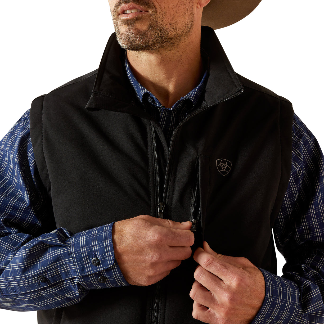 Man wearing a black vest over a plaid shirt with a visible brand logo.