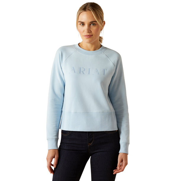Woman wearing a light blue sweatshirt with 'Ariat' branding on a white background