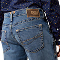 Close-up of blue jeans with Ariat denim label, worn by a person in a patterned shirt.
