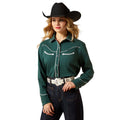 Woman wearing a green cowboy shirt with white stitching, black cowboy hat, and dark jeans on a beige background.