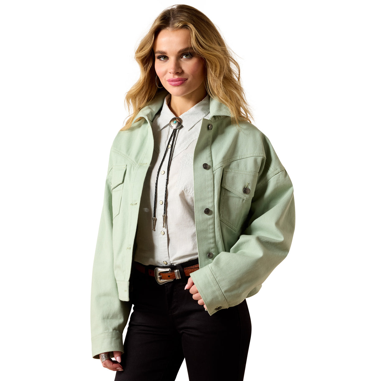A woman modeling a light sage green cropped utility jacket over a white collared shirt with a tie, paired with black pants and a brown belt, on a solid white background.