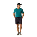 A man wearing a teal short-sleeve athletic t-shirt, dark charcoal drawstring shorts, a black baseball cap, and black sneakers, standing against a solid white background.