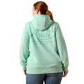 A woman viewed from the back wearing a mint green hoodie with a large 'Ariat' logo printed on the back, paired with blue jeans against a plain white background.