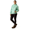 A woman modeling a light mint green hooded sweatshirt with black leggings and brown boots, standing against a plain white studio background.