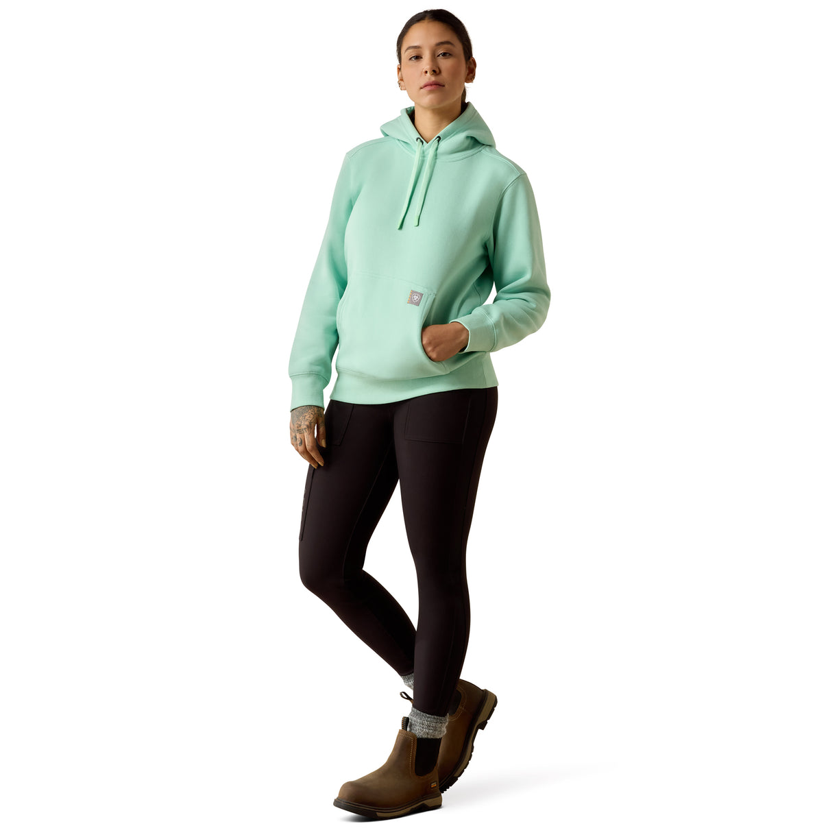 A woman modeling a light mint green hooded sweatshirt with black leggings and brown boots, standing against a plain white studio background.