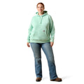 A woman modeling a mint green pullover hoodie with drawstrings and a kangaroo pocket, paired with blue jeans and brown shoes, against a plain white background.