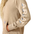 Close-up of a person wearing a heather tan pullover hoodie with a large vertical ARIAT logo printed on the sleeve in cream and light blue, plus a small brand patch on the pocket.