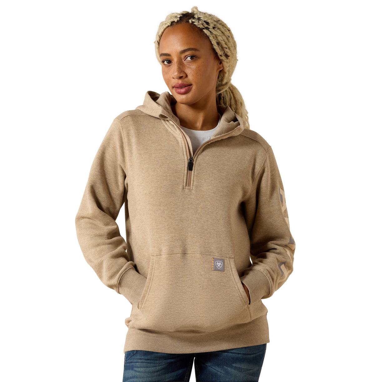 A woman wearing a light tan heathered pullover hoodie with a half-zip neckline, kangaroo pocket, and a small square logo patch near the lower front, paired with blue jeans against a plain white background.