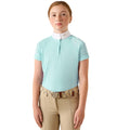A young girl wearing a light blue equestrian polo shirt with a white collar, paired with beige riding pants and a patterned belt