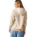 A woman shown from the back wearing a light beige hooded sweatshirt and blue jeans against a plain white background.