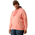 A woman modeling a coral pink hooded sweatshirt with red drawstrings and red vertical branding on the sleeve, paired with blue jeans and a brown cap, against a plain white background.