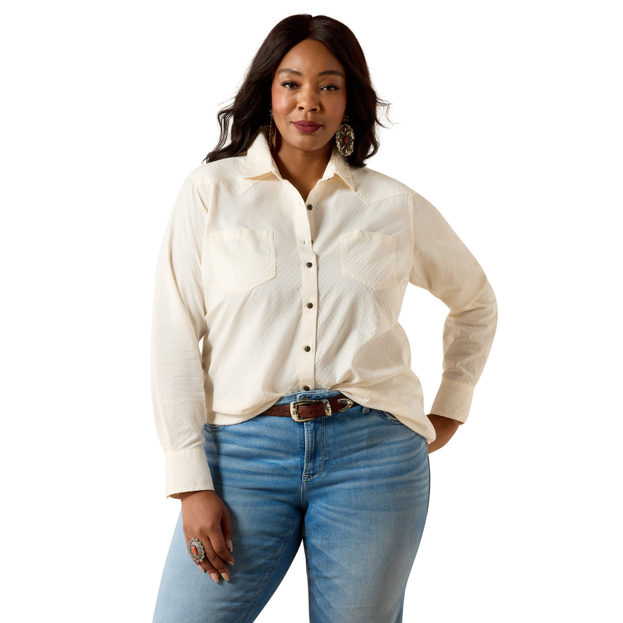 A woman modeling a cream-colored long-sleeve western-style button-up shirt with two chest pockets, paired with blue jeans and a brown belt, against a solid white background.