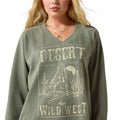 A woman wearing a sage green V-neck sweatshirt with a distressed desert graphic that reads 'DESERT WILD WEST' and 'FEEL THE SUNSET'.