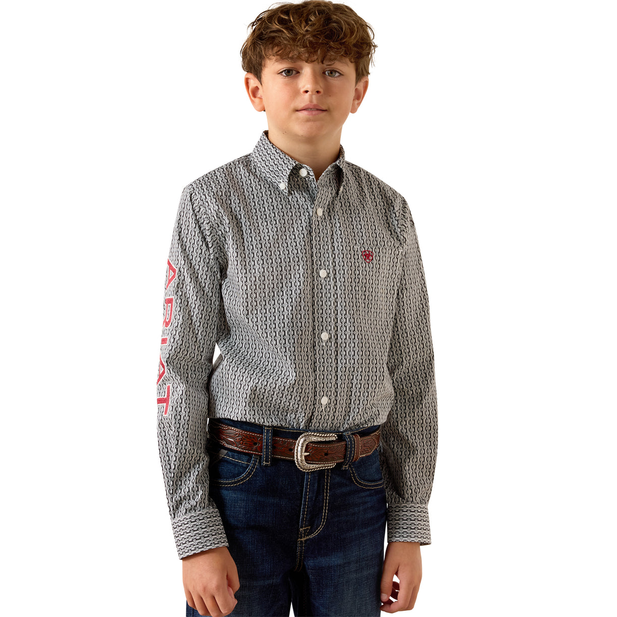 A young boy modeling a long-sleeve button-down shirt with a gray and white geometric pattern, featuring an Ariat logo on the left chest and a red graphic on the right sleeve, paired with dark blue jeans and a brown belt.