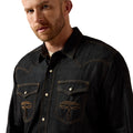 A close-up of a man wearing a dark indigo denim western shirt with contrast tan stitching and embroidered chest details, set against a plain beige background.
