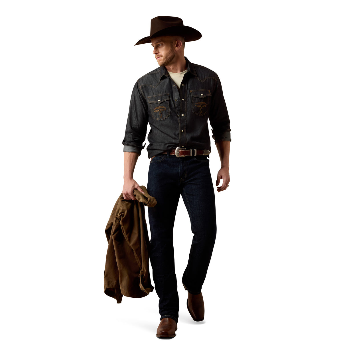 A man wearing a dark gray denim western shirt, dark blue jeans, a brown cowboy hat, and brown boots, holding a brown jacket in one hand against a plain white background.