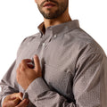 A close-up of a man wearing a brown long-sleeve button-down shirt with a small white geometric pattern, adjusting the cuff with one hand.