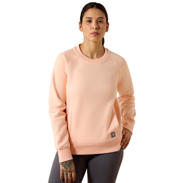 A woman modeling a light peach crewneck sweatshirt with raglan sleeves and a small black brand patch near the lower hem, paired with dark gray leggings against a plain white background.