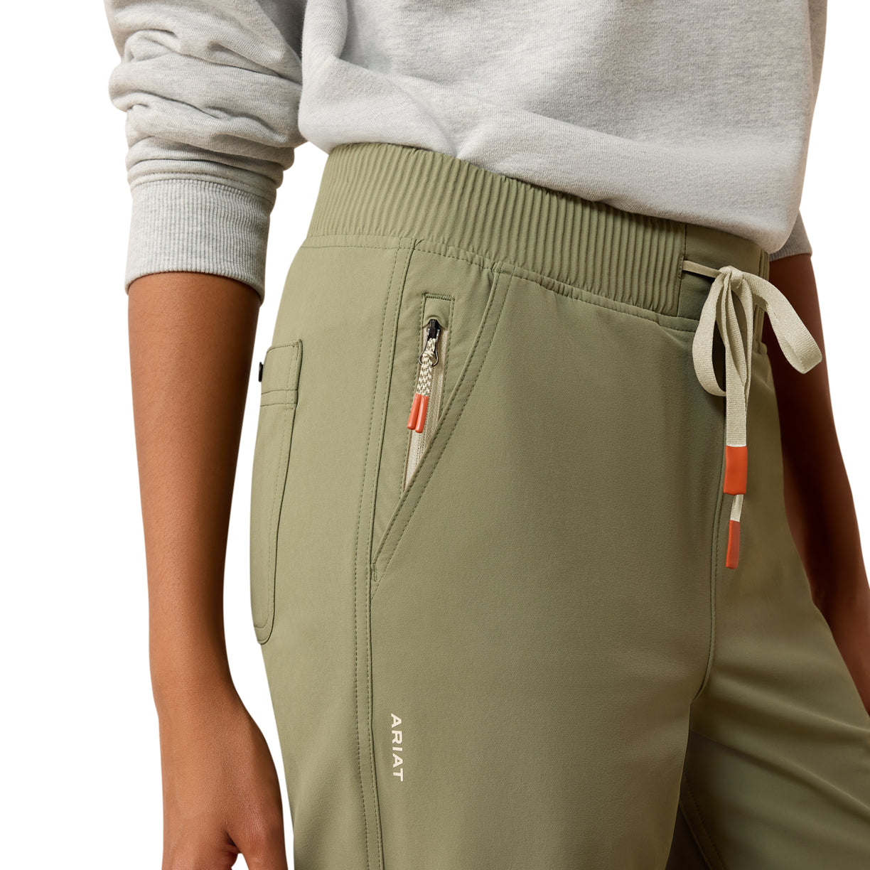 Person wearing olive green pants with drawstring and zippers on a plain background