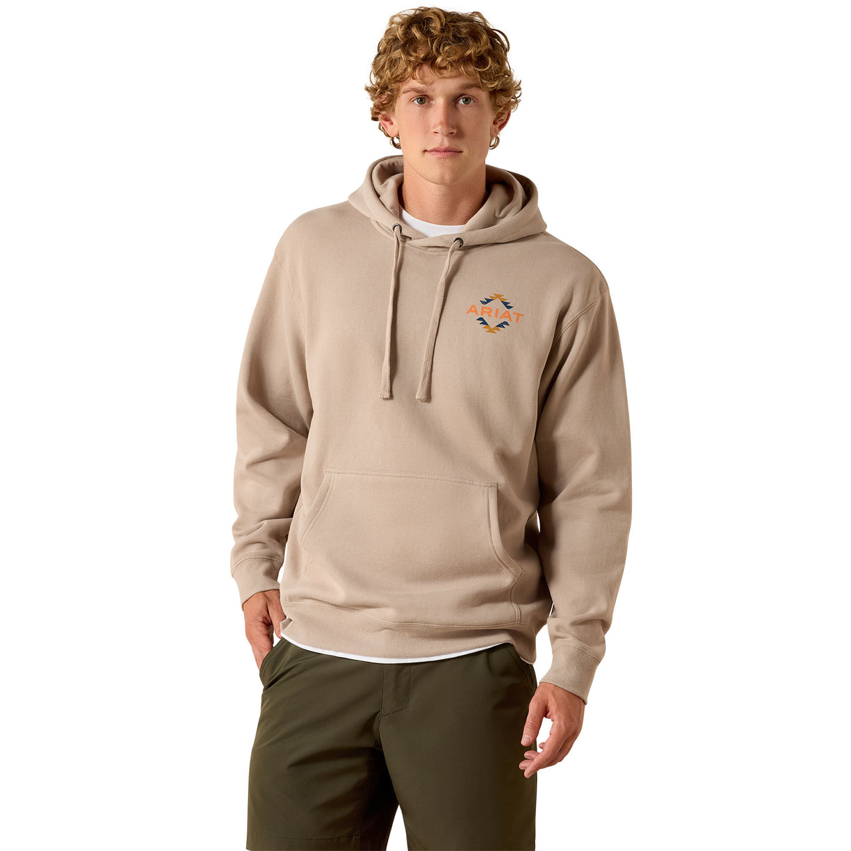 A man wearing a beige pullover hoodie with a small orange and blue mountain logo on the left chest, paired with dark olive pants, against a plain white background.