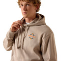 A young man with curly blond hair wearing a beige pullover hoodie with a small orange and blue ARIAT logo on the left chest, posed against a solid white background.