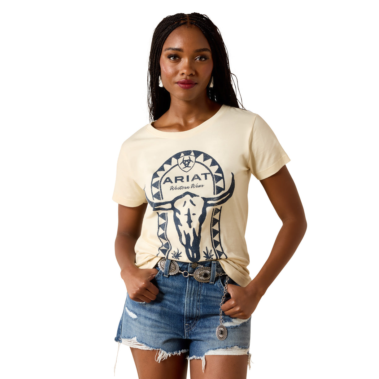 A woman modeling a cream-colored short-sleeve t-shirt with a dark blue graphic of a skull and the text 'ARIAT Outfitter' on the front, paired with distressed denim shorts against a plain white background.