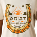 A close-up of a person wearing a white T-shirt with a large graphic print on the chest. The design shows a horseshoe outline with orange and beige sections, black text reading 'ARIAT BOOT and DENIM COMPANY,' and '1993' at the bottom, plus small star and mountain motifs.
