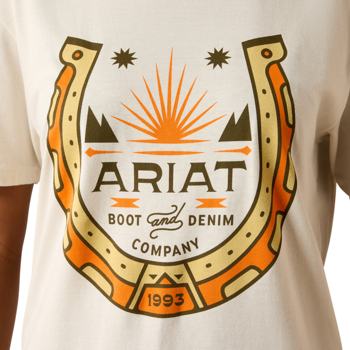 A close-up of a person wearing a white T-shirt with a large graphic print on the chest. The design shows a horseshoe outline with orange and beige sections, black text reading 'ARIAT BOOT and DENIM COMPANY,' and '1993' at the bottom, plus small star and mountain motifs.