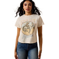 Model wearing a beige Ariat t-shirt with a graphic design and text, standing 