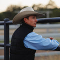 Man wearing a cowboy hat and black vest standing behind a fence with trees in the background