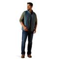 Man wearing a blue vest over a green shirt with dark jeans on a white background
