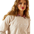 Woman wearing a beige sweater with 'Ariat' embroidery on a white background
