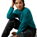 Woman wearing a teal long-sleeve top and dark patterned pants on a white background