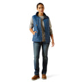 Woman wearing a blue vest over a gray shirt with jeans on a white background