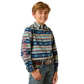 A young boy smiling and wearing a long-sleeve western button-down shirt with a colorful geometric pattern in navy blue, orange, and beige, paired with jeans and a belt.