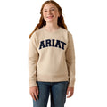 A young woman with long light brown hair smiling, wearing a beige crewneck sweatshirt with 'ARIAT' printed in navy blue across the chest, paired with blue jeans, against a plain white background.
