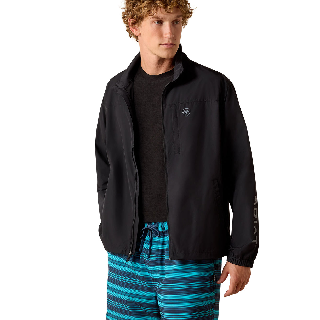A man wearing a black lightweight zip-up jacket with a small shield logo on the left chest, a dark crewneck shirt, and blue and black striped shorts, photographed against a plain white background.