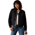 A woman modeling a black denim-style jacket with silver snap and button details, worn over a white top and blue jeans against a plain white background.