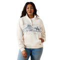 A woman modeling a cream-colored quarter-zip hoodie with a blue desert landscape graphic on the front, paired with blue jeans, against a plain white background.