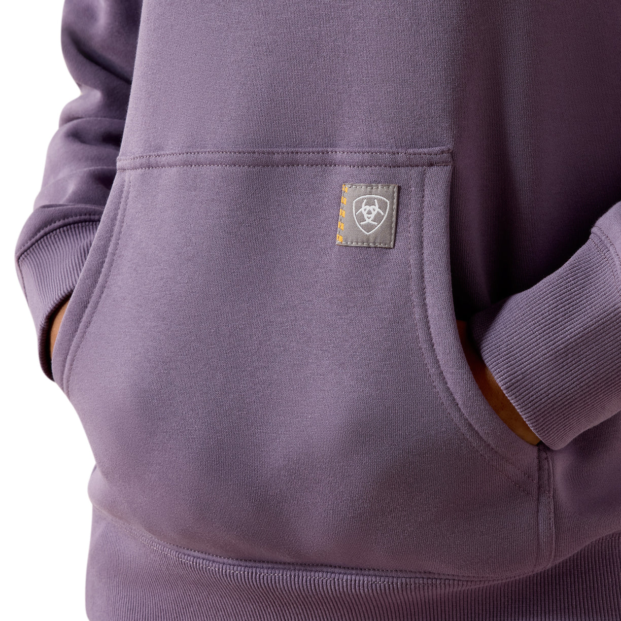 Close-up of a person wearing a dusty purple hoodie, showing the front pocket with a small shield logo patch on the chest.